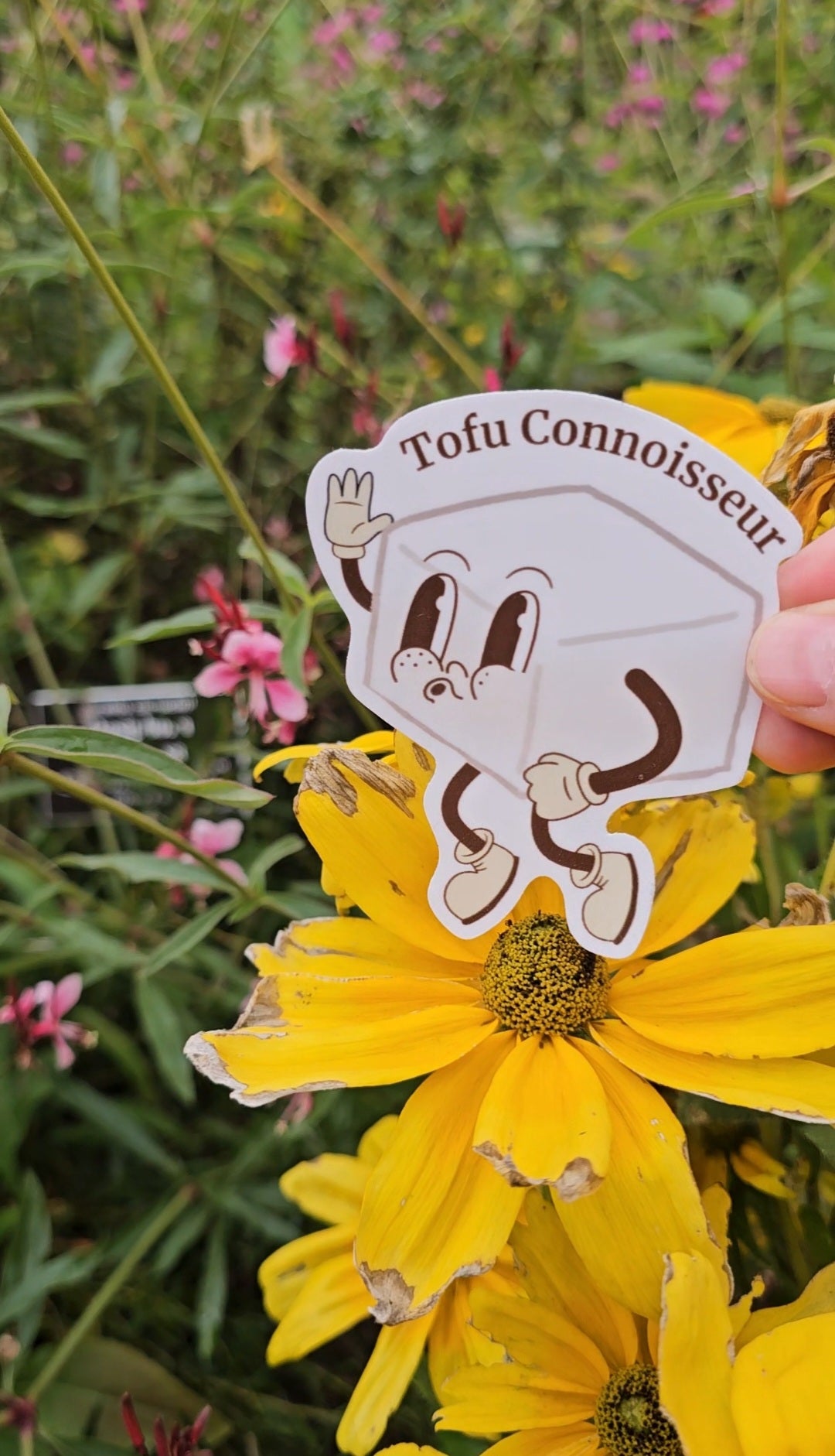 Hand holding a 'Tofu Connoisseur' sticker over a yellow flower with a blurred natural background.