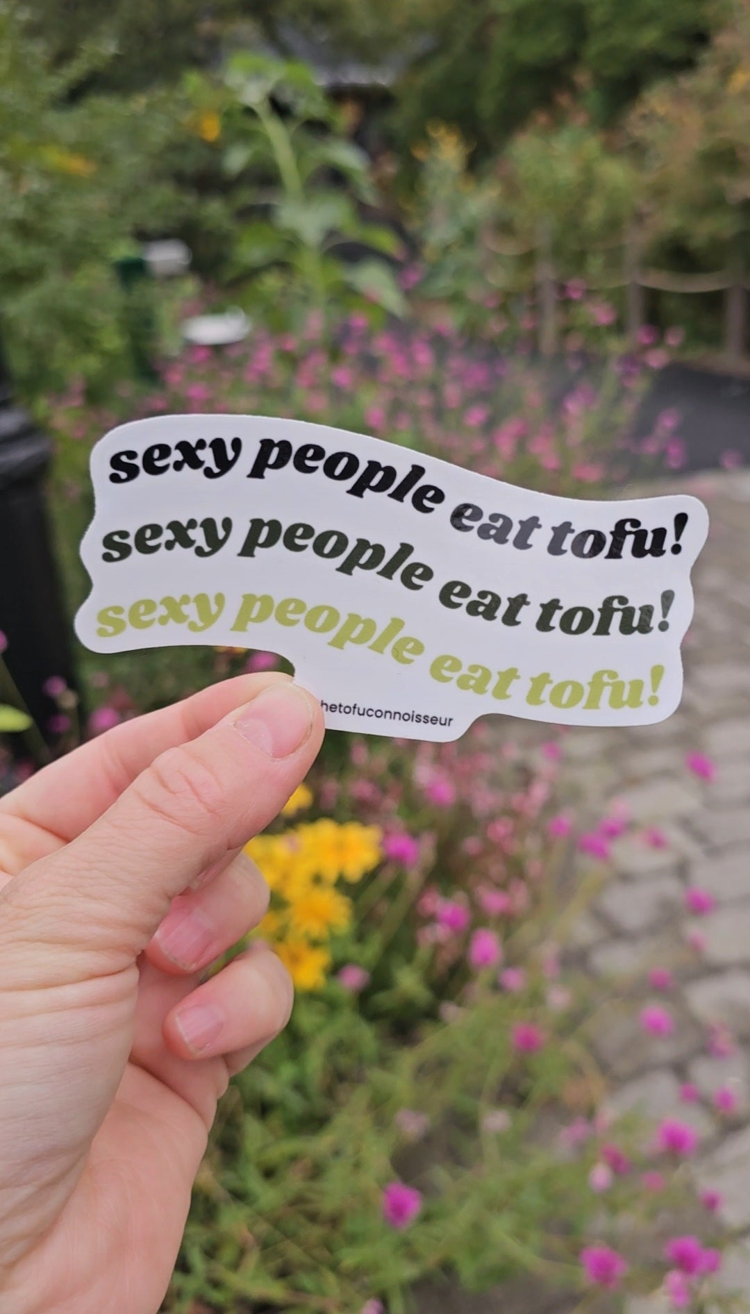 Hand holding a sticker with 'sexy people eat tofu!' text against a blurred natural background